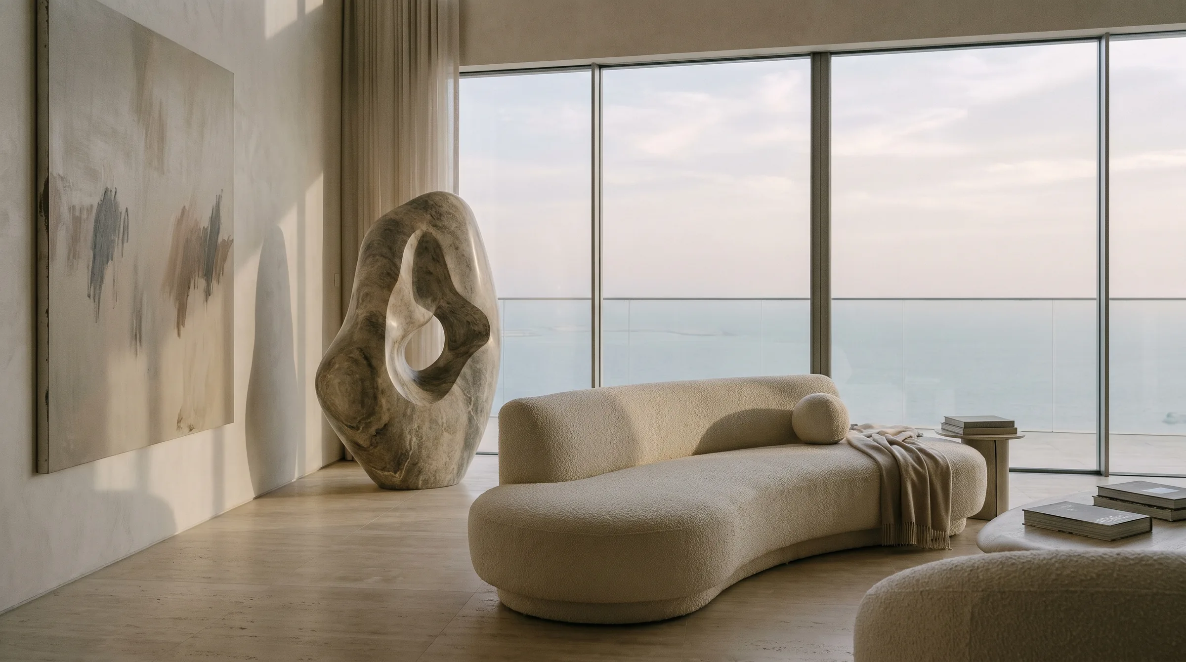 A double-height penthouse with travertine floors, a curved cream sofa, a stone sculpture and a wide view of the sea
